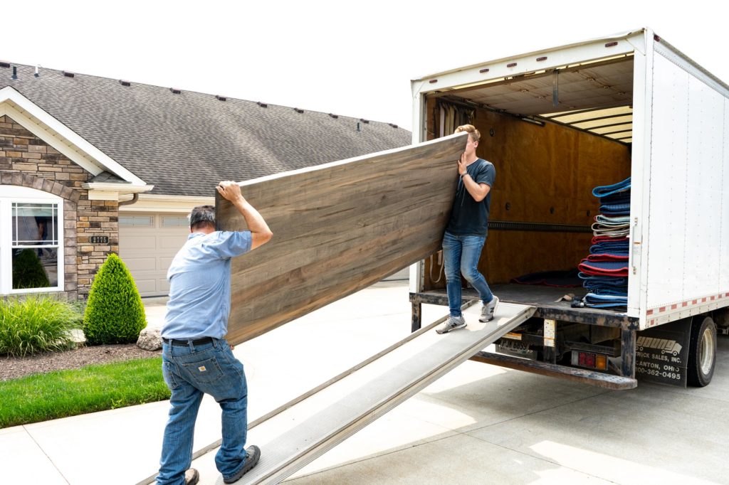 Two men unloading a large wooden furniture piece from a moving truck in front of a suburban house.