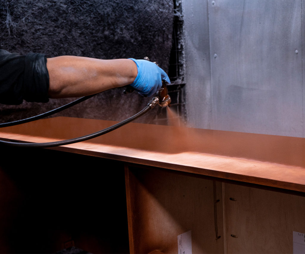 Woodworker applying a finish to a wooden surface using a spray gun, wearing blue gloves.