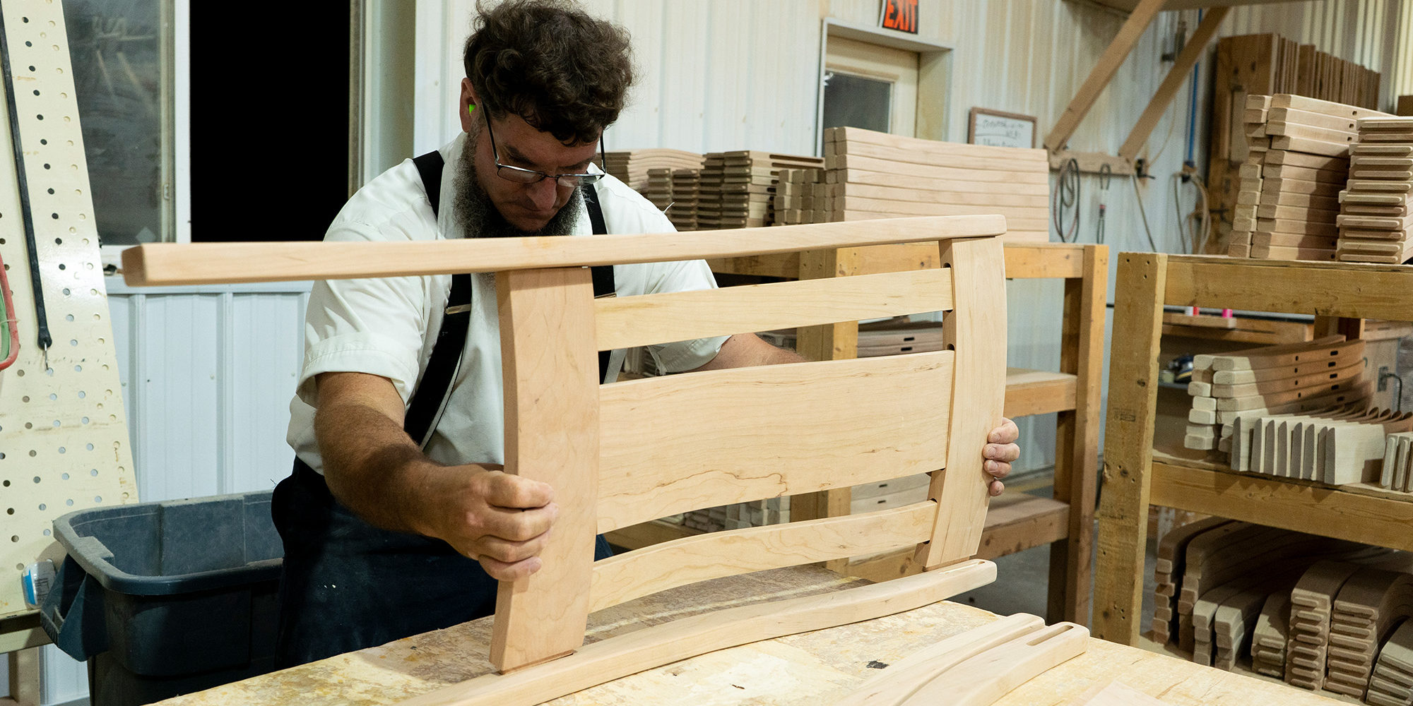 Craftsman in a workshop assembling a wooden chair backrest, wearing suspenders and glasses.