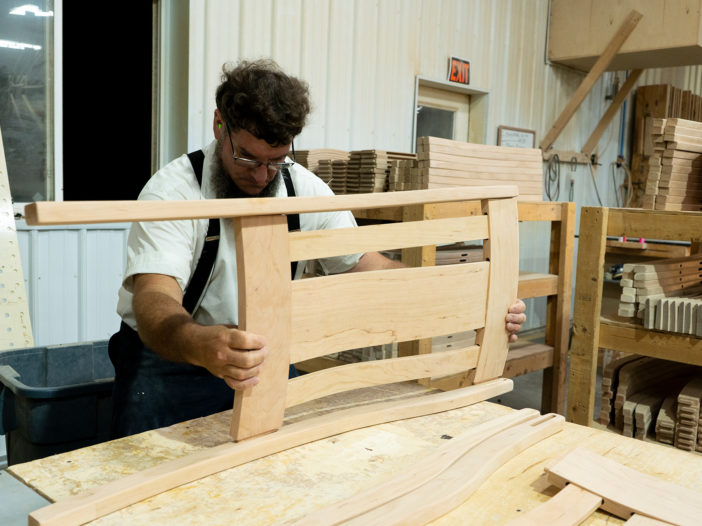 Craftsman in a workshop assembling a wooden chair backrest, wearing suspenders and glasses.